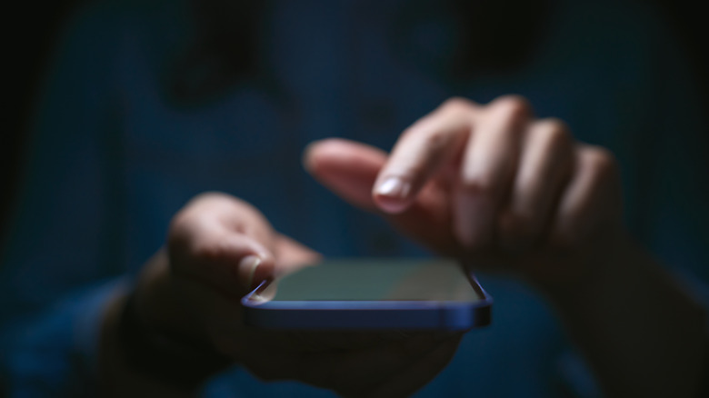 Two hands holding a smartphone in a dark environment, suggesting a malicious user.