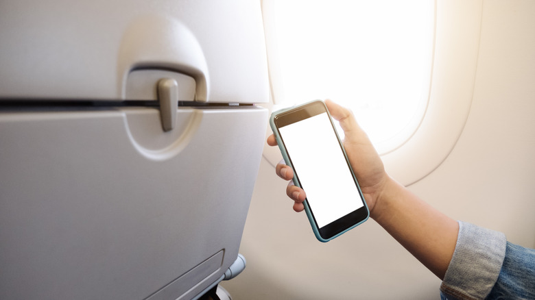 A person holding a smartphone near an airplane seatback in a cabin.