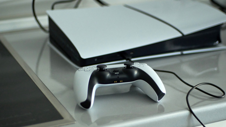 A PS5 DualSense controller resting on a table next to the console with a charging cable.