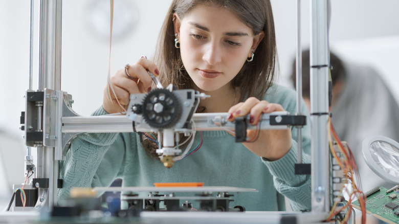 A person leaning in to inspect the print head and gantry of an FDM 3D printer.