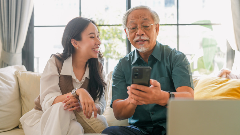 An elderly man sitting on a couch smiling while using a smartphone with a young woman looking on.