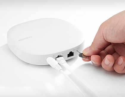 A close-up of a person using a pin tool to press the reset button on a SmartThings Wi-Fi hub.