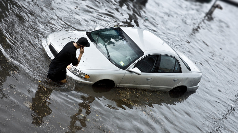 A man looking distressed while standing next to his car submerged in floodwaters.