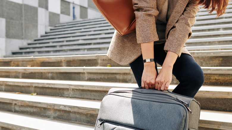 A woman struggling to pull a large, heavy suitcase up a steep flight of concrete stairs.