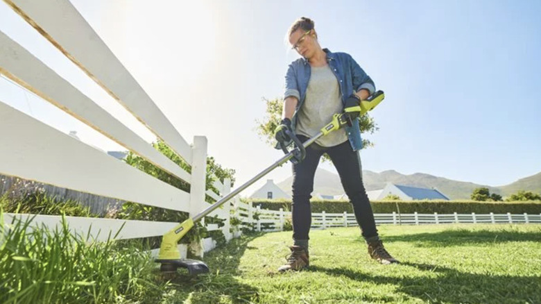 A woman using a Ryobi cordless string trimmer to edge a lawn along a white fence.