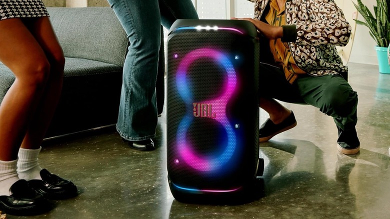 A JBL PartyBox speaker on the floor with people dancing and colorful lights glowing.